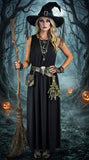 A woman modeling a black witch costume dress with a matching pointed witch hat, holding a broomstick in a dark, foggy forest setting with glowing jack-o'-lanterns.